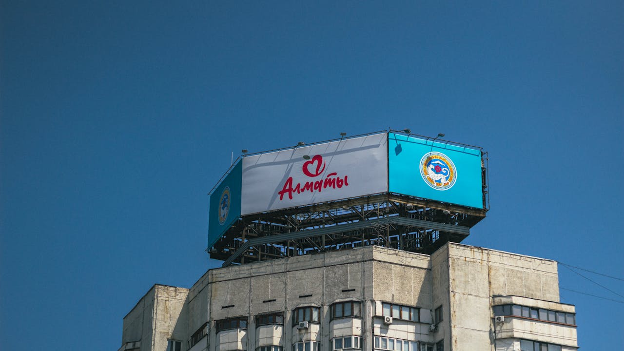 High-rise building with Almaty billboard and cityscape under clear blue daytime sky.